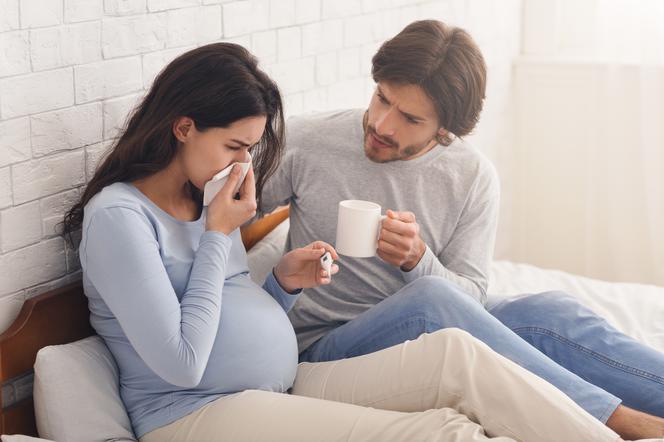 Przeziębiona kobieta w ciąży leży w łóżku. Partner podaje jej kubek z gorącą herbatą.