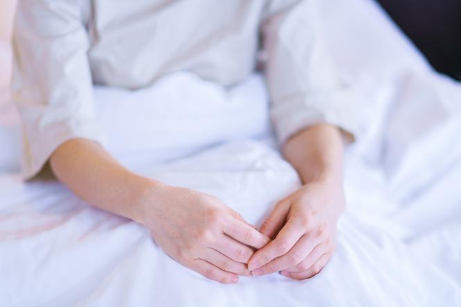 Kobieta w średnim wieku siedzi owiniętą białą pościelą na łóżku.